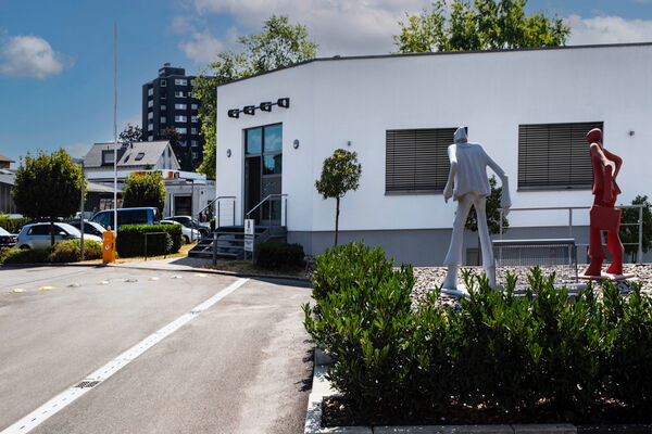Flacher weißer Bürobau mit Eingang, davor zwei überlebensgroße Figurenskulpturen zwischen Hecken