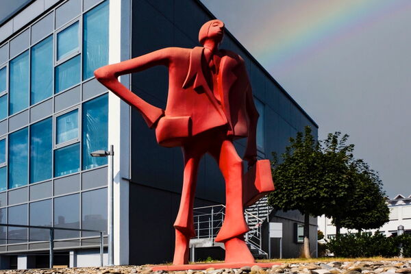 Rote Figurenskulptur vor einem verglasten Bürogebäude, dahinter ein Regenbogen