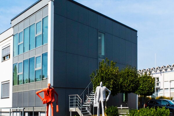 Anthrazitfarbener Bürokubus mit Glasfront, davor eine rote und eine weiße Figurenskulptur