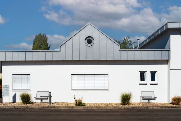 Weißes Gebäude mit Zinkdach und rundem Fenster im Giebel, davor zwei Bänke